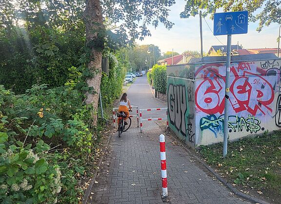 Zu sehen ist der Verbindungsweg zwischen Zamenhofweg und Siedlerweg. Auf dem Weg ist sowohl ein Steckpfosten (Poller) als auch eine Umlaufsperre (Drängelgitter) angebracht.