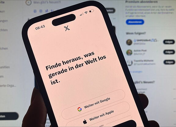 Ein Smartphone mit der geöffneten App "X" vor einem Computer-Bildschirm