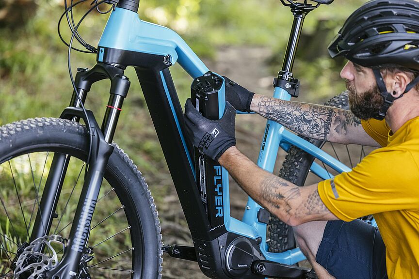 Pedelec-Akku wird entnommen Ein Mann in gelbem Trikot und schwarzem Fahrradhelm entnimmt einen schwarzen Akku aus dem hellblauen Rahmen seines Pedelecs.